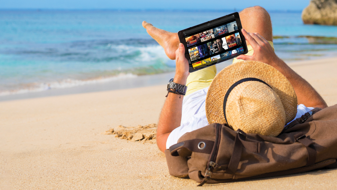 A person uses a tablet as they lie on a beach.