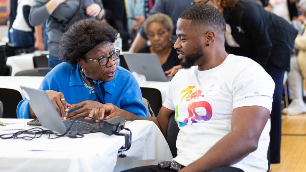 The image depicts two individuals seated at a table, working on a laptop. They are in a room with other people, possibly during an event or meeting. One person is wearing a blue top and the other a white t-shirt with colorful text and graphics.